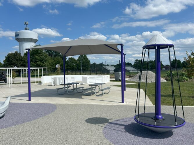 4 post fabric shade structure with 2 gray picnic tables next to a multi-person spinner