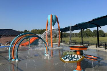 blue and orange splash pad with multiple water play events with blue shade above benches for seating