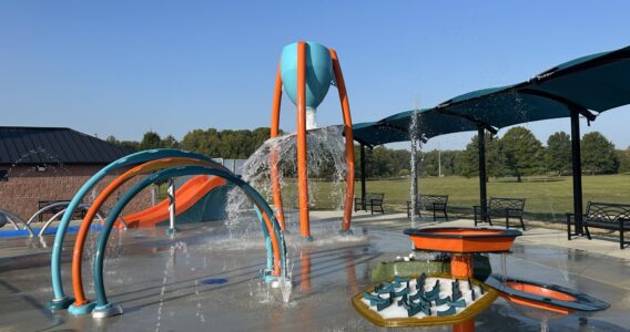 blue and orange splash pad with multiple water play events with blue shade above benches for seating